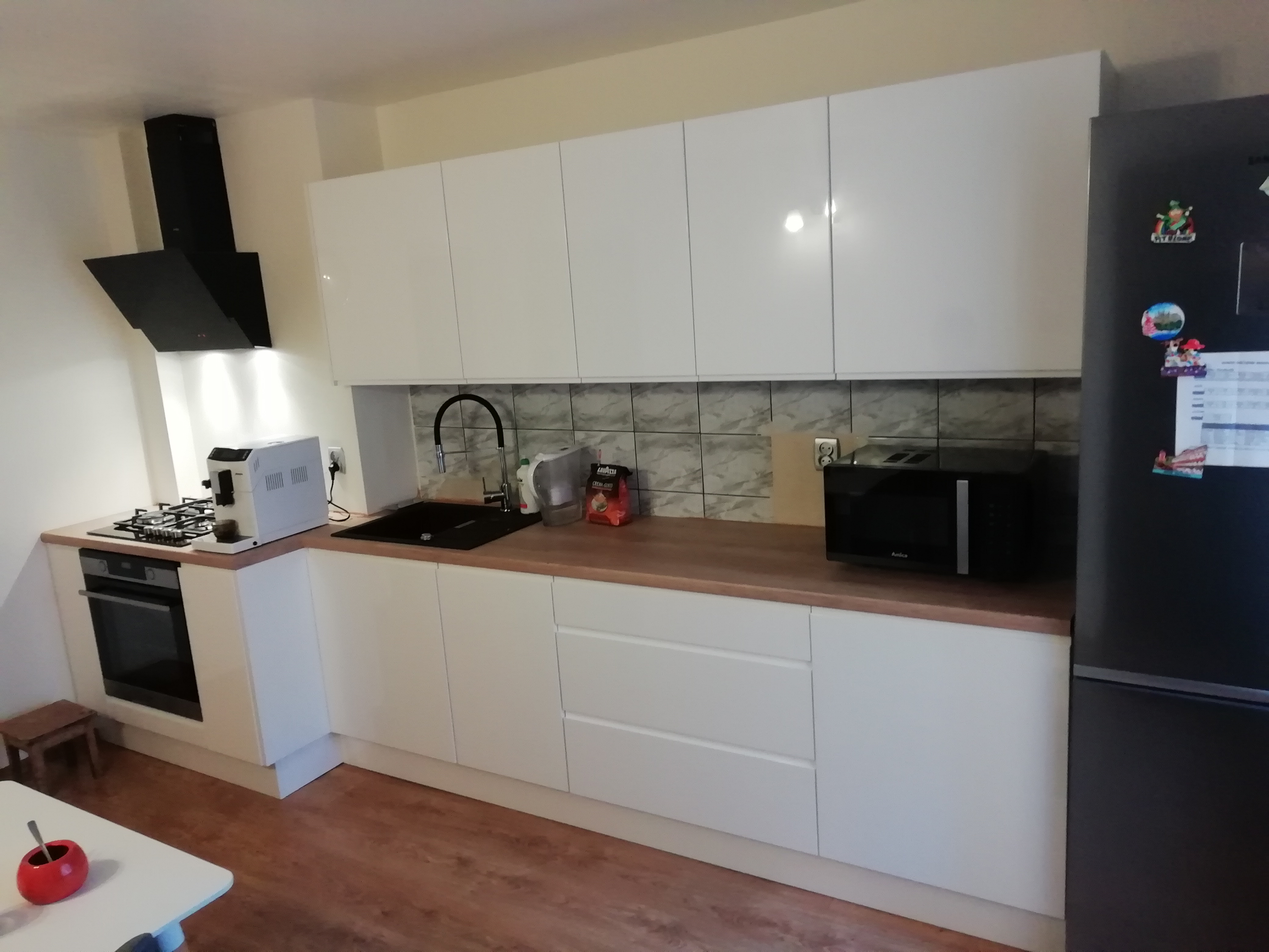 Nowoczesna, jasna kuchnia z białymi, błyszczącymi frontami szafek, drewnianym blatem, czarnym okapem i czarnym zlewem. Na blacie widoczny ekspres do kawy, mikrofalówka i akcesoria kuchenne.