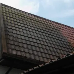 Częściowo wyczyszczony dach z metalowej dachówki, z widoczną różnicą w kolorze między umytą a zabrudzoną powierzchnią, na tle błękitnego nieba.