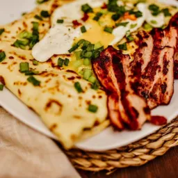 Naleśniki z jajkiem sadzonym, boczkiem i posiekanym szczypiorkiem na białym talerzu, ułożone na plecionym podkładce na drewnianym stole.