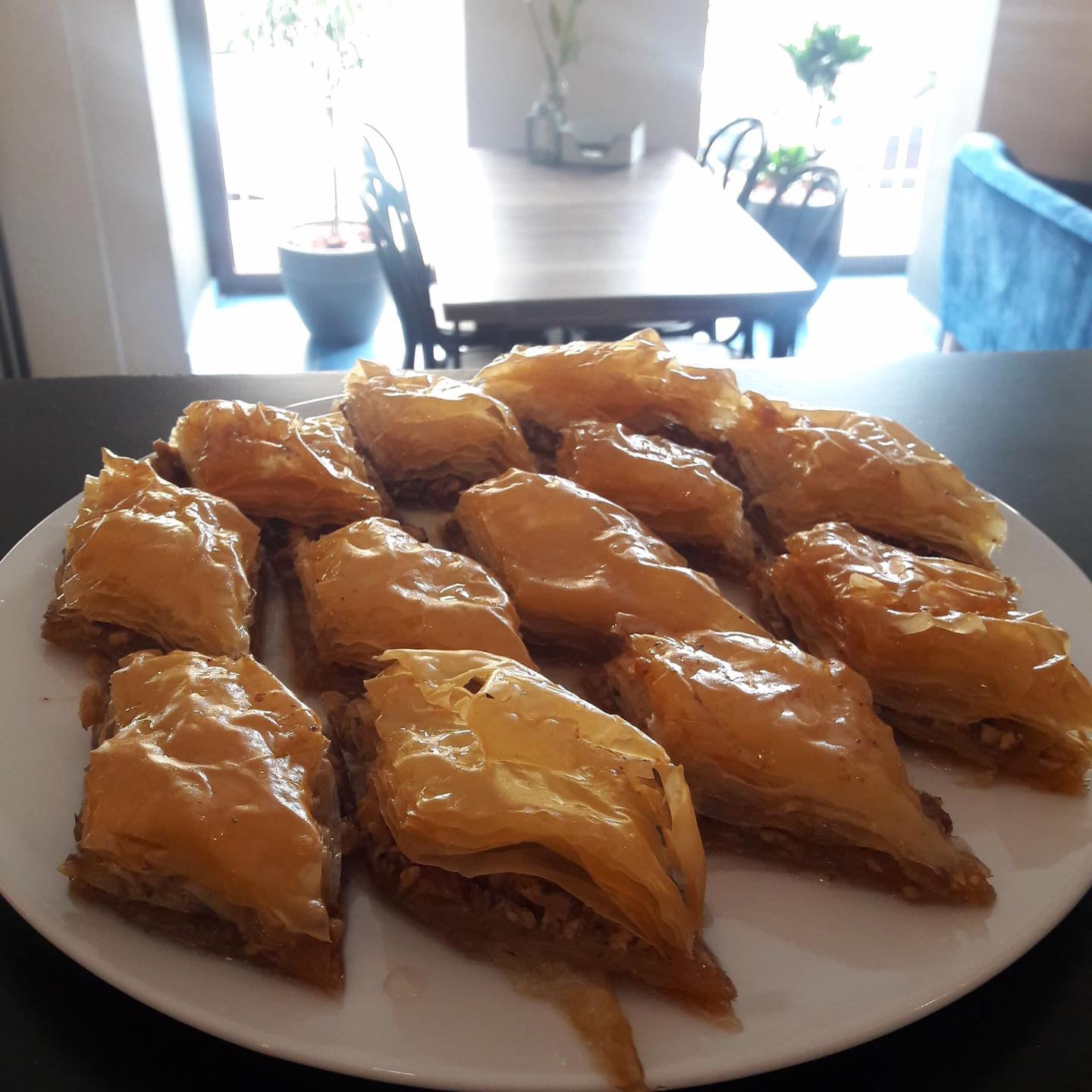 Taca z dwunastoma kawałkami baklawy, polanej syropem, na białym talerzu, z widocznym stolikiem i krzesłami w tle.