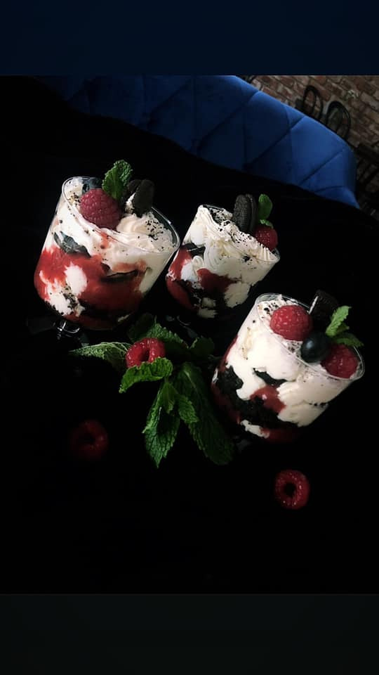 Trzy desery warstwowe w przezroczystych pucharkach, z bitą śmietaną, sosem malinowym, pokruszonymi ciastkami Oreo, malinami i listkami mięty na czarnym tle. W tle widoczny fragment niebieskiej sofy...