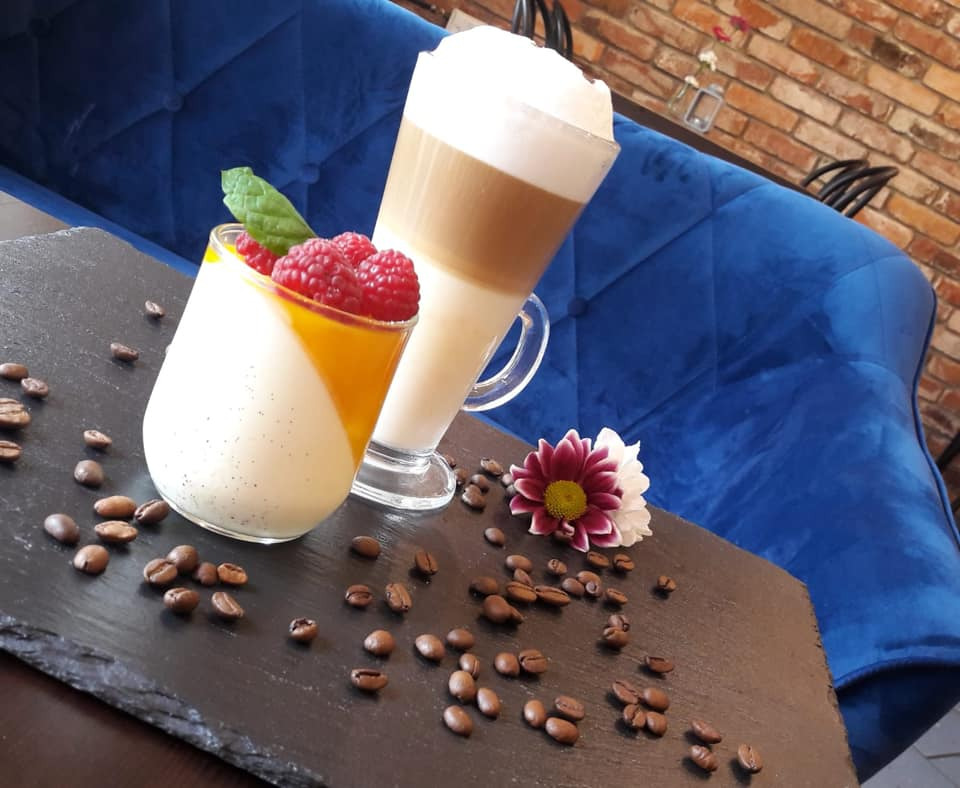Elegancko podane latte i deser panna cotta z malinami i listkiem mięty, otoczone ziarnami kawy na łupkowym talerzu, z kwiatem w tle i niebieskim obiciem kanapy.