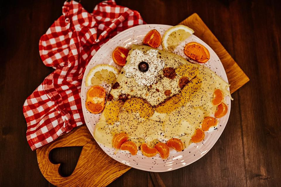 Delikatny naleśnik na różowym talerzu, udekorowany mandarynkami, pomarańczami i cytrynami, posypany przyprawami, z kleksem bitej śmietany i pojedynczą jagodą na wierzchu, leżący na drewnianej desce...