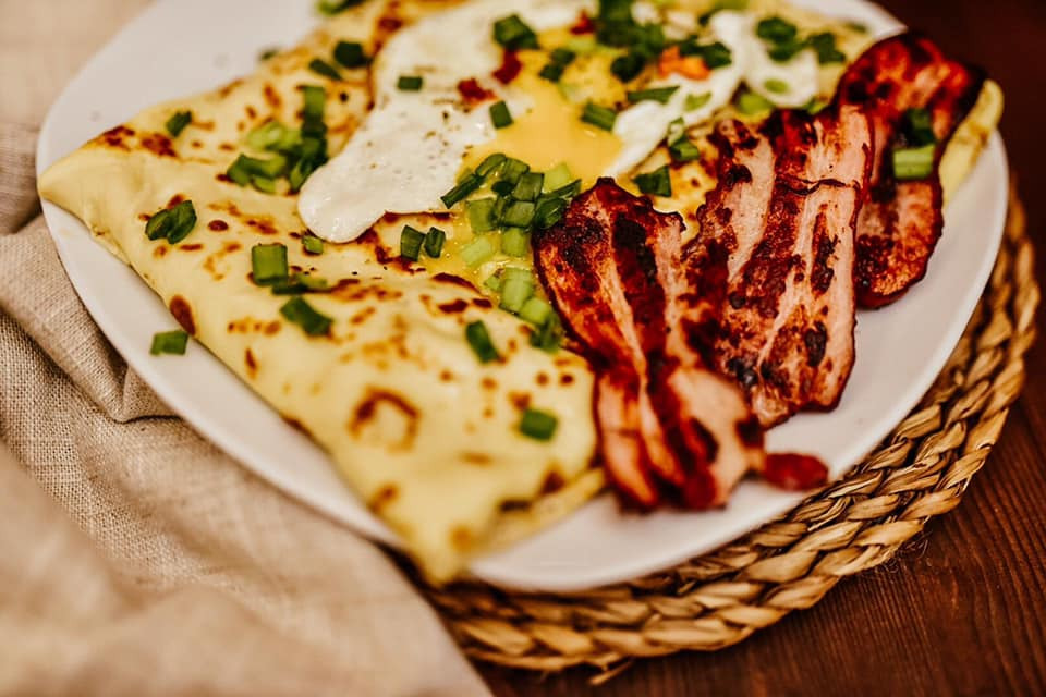 Naleśniki z jajkiem sadzonym, boczkiem i posiekanym szczypiorkiem na białym talerzu, ułożone na plecionym podkładce na drewnianym stole.