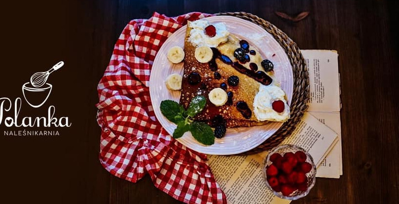 Naleśnik z owocami i bitą śmietaną na talerzu z dekoracją z książek i kratowanej ściereczki, obok logo naleśnikarni.