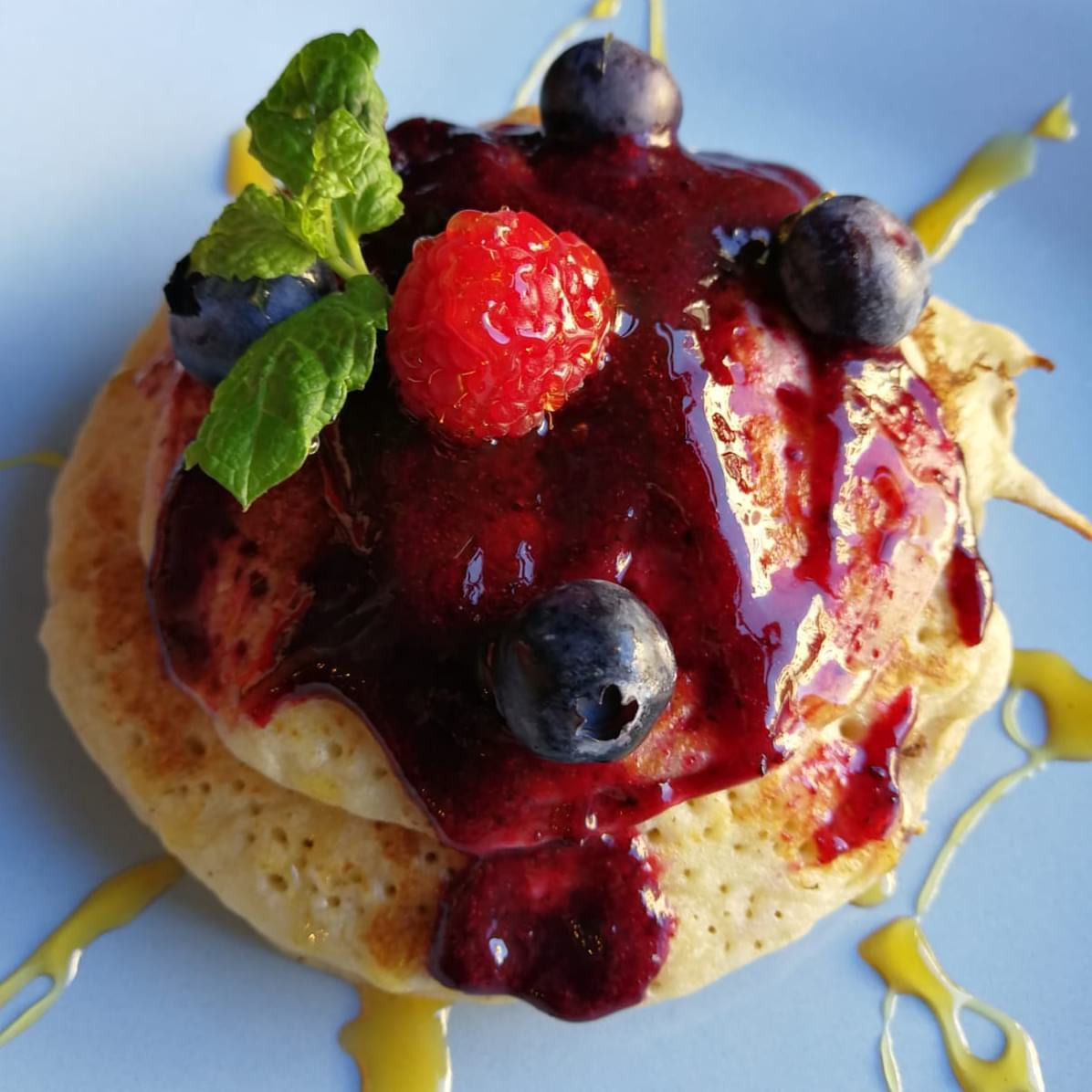 Dwa puszyste naleśniki polane gęstym sosem jagodowym, ozdobione świeżą maliną, borówkami i listkiem mięty, całość na jasnoniebieskim talerzu z dodatkiem syropu klonowego.