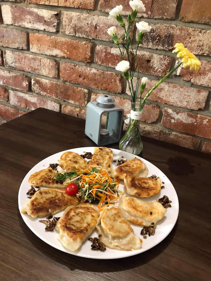 Rząd pierogów gyoza na białym talerzu, ozdobiony sałatką z marchewki i kiełków, z pomidorkiem cherry, ustawiony na ciemnym drewnianym stole z dekoracją kwiatową i ceglaną ścianą w tle.