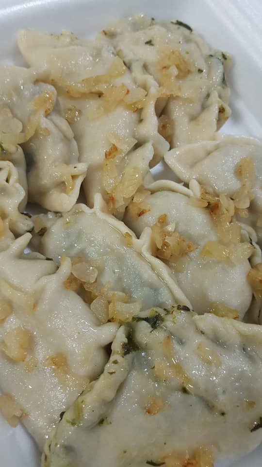 Bliski kadr na porcję gotowanych pierogów z farszem szpinakowym, polanych podsmażoną cebulką w plastikowym pojemniku.