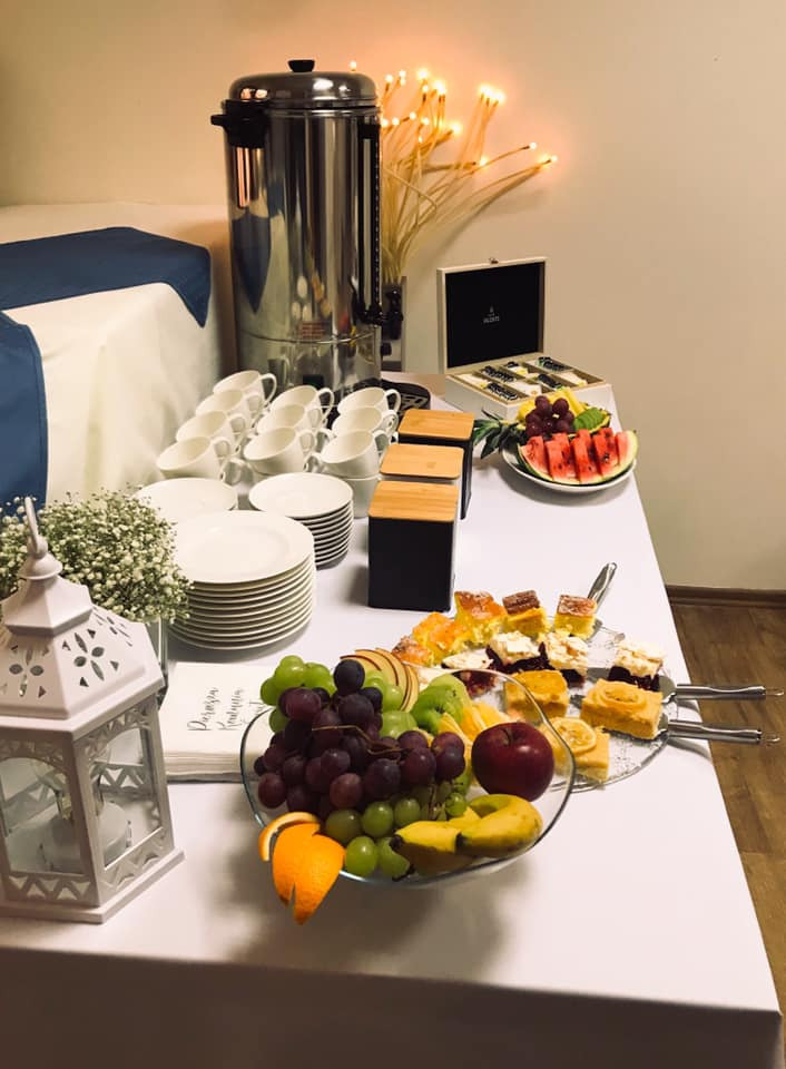 Stół cateringowy z dyspensorem do kawy, piramidą talerzy i filiżanek, paterą owoców i ciast, ozdobnym lampionem i serwetkami z napisem.