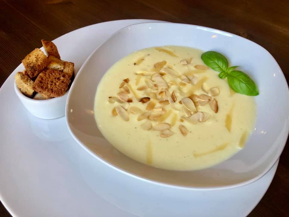 Kremowa zupa z migdałami i bazylią w białym, asymetrycznym talerzu, obok miseczka z grzankami, na drewnianym stole.