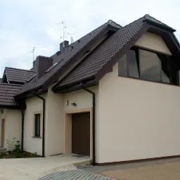 Jasna elewacja domu jednorodzinnego z brązowym dachem i stolarką okienną, widok z boku na podjazd i fragment ogrodu