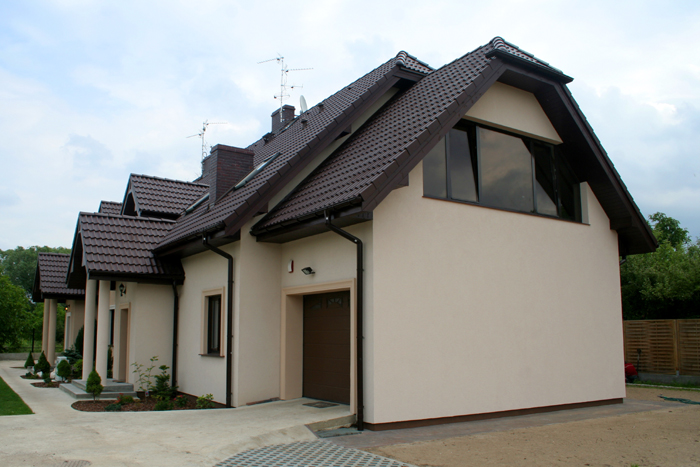 Jasna elewacja domu jednorodzinnego z brązowym dachem i stolarką okienną, widok z boku na podjazd i fragment ogrodu