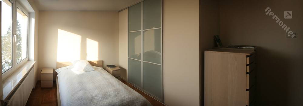 Jasny pokój z łóżkiem, przesuwną szafą z mlecznego szkła i komodą w odcieniach beżu, oświetlony porannym słońcem wpadającym przez okno.