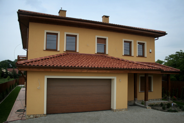 Ocieplona elewacja dwukondygnacyjnego domu z brązową bramą garażową, czerwoną dachówką i oknami w brązowych ramach.