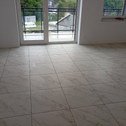 MAL-FLIZ - Jasne, marmurowe płytki podłogowe w nowym wnętrzu z oknami balkonowymi. Widok na zewnątrz, gdzie widać zieleń i zabudowania. Fragment drabiny w prawym rogu.