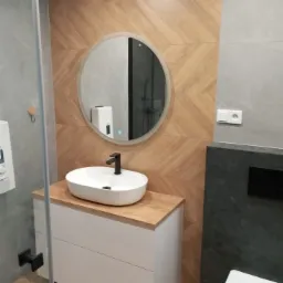 Nowoczesna łazienka z umywalką nablatową, okrągłym lustrem z podświetleniem, i drewnopodobnymi płytkami ułożonymi w jodełkę na ścianie.