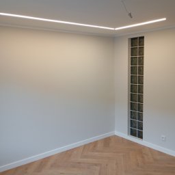 MAL-FLIZ - Jasne wnętrze po remoncie: parkiet w jodełkę, szara ściana z oknem z luksferów i listwy przypodłogowe. Oświetlenie LED na suficie.