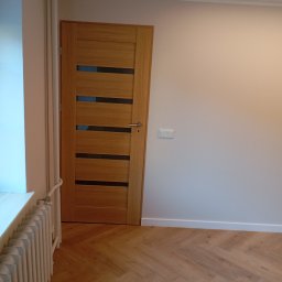 MAL-FLIZ - Jasne panele ułożone w jodełkę, drewniane drzwi z przeszkleniami i biała ściana w Urbanowicach. Widoczny grzejnik i listwa przypodłogowa.