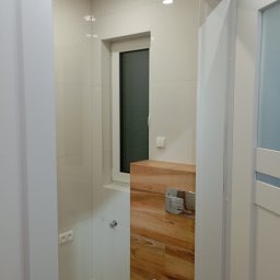 MAL-FLIZ - Nowoczesna łazienka z białymi płytkami i akcentem drewnianym. Wisząca toaleta z zabudową. Okno w białej ramie. Widok zza drzwi.