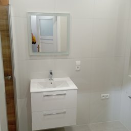 MAL-FLIZ - Biała umywalka z szafką i lustrem w nowoczesnej łazience z białymi płytkami. Widoczne gniazdka i fragment kabiny prysznicowej z drewnianym akcentem.