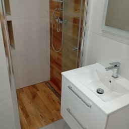 MAL-FLIZ - Nowoczesna łazienka z drewnopodobną podłogą i kabiną prysznicową typu walk-in. Biała szafka z umywalką i chromowaną armaturą. Minimalistyczny design, jasne płytki ścienne.