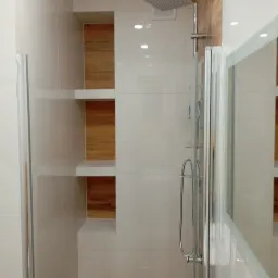 Nowoczesna kabina prysznicowa z przeszklonymi drzwiami i drewnianymi wnękami w jasnych płytkach, minimalistyczny design, chromowane wykończenia.