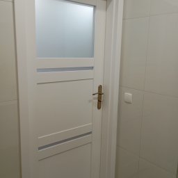 MAL-FLIZ - Białe drzwi z szybą i złotą klamką w łazience wyłożonej jasnymi, kwadratowymi płytkami. Widoczny włącznik światła.