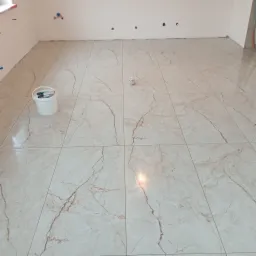 Jasne, marmurowe płytki podłogowe w nowym pomieszczeniu. Widoczny biały pojemnik z narzędziami i wystające kable. Ściany przygotowane do dalszych prac.