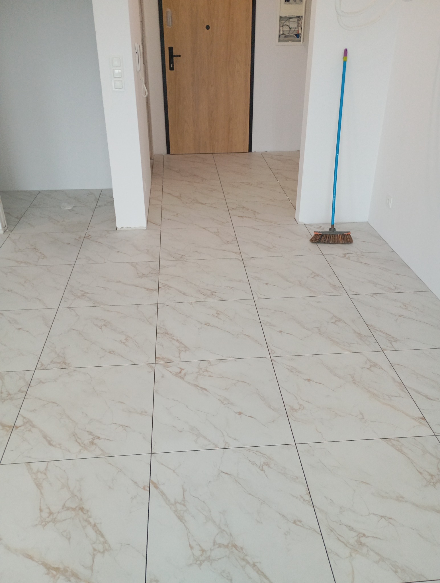 Jasne, marmurowe płytki podłogowe w korytarzu z drewnianymi drzwiami. W rogu stoi niebieska zmiotka. Widok na wykończone podłogi po remoncie.