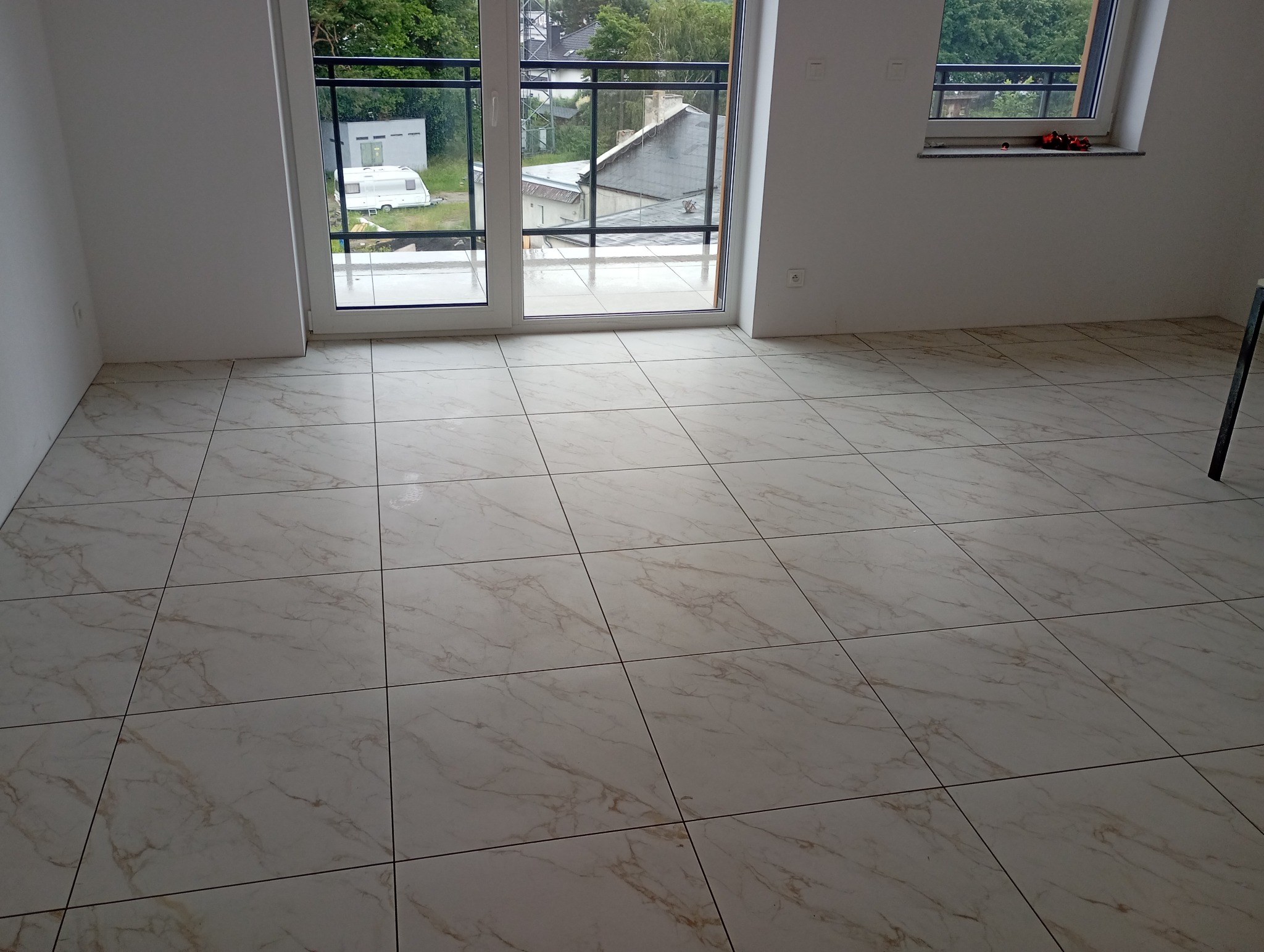 Jasne, marmurowe płytki podłogowe w nowym wnętrzu z oknami balkonowymi. Widok na zewnątrz, gdzie widać zieleń i zabudowania. Fragment drabiny w prawym rogu.