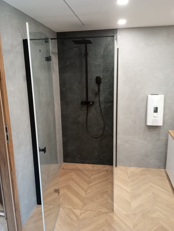 Nowoczesna kabina prysznicowa z czarną armaturą na tle szarej ściany i drewnopodobnej podłogi ułożonej w jodełkę, widoczna z boku.