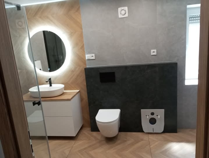 Nowoczesna łazienka z okrągłym lustrem LED, białą umywalką nablatową i podwieszanym WC. Płytki imitujące drewno na podłodze i ścianie za umywalką.