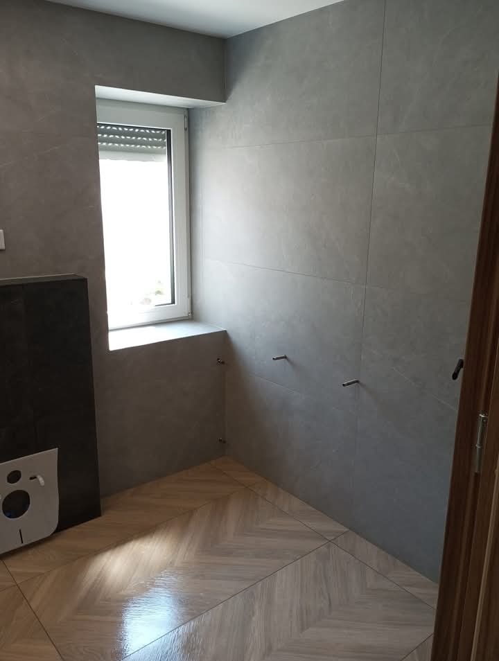 Nowoczesna łazienka z szarymi płytkami ściennymi i podłogą ułożoną w jodełkę. Widoczne metalowe elementy armatury i okno z roletą. Minimalistyczny design.