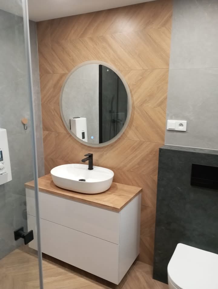 Nowoczesna łazienka z umywalką nablatową, okrągłym lustrem z podświetleniem, i drewnopodobnymi płytkami ułożonymi w jodełkę na ścianie.