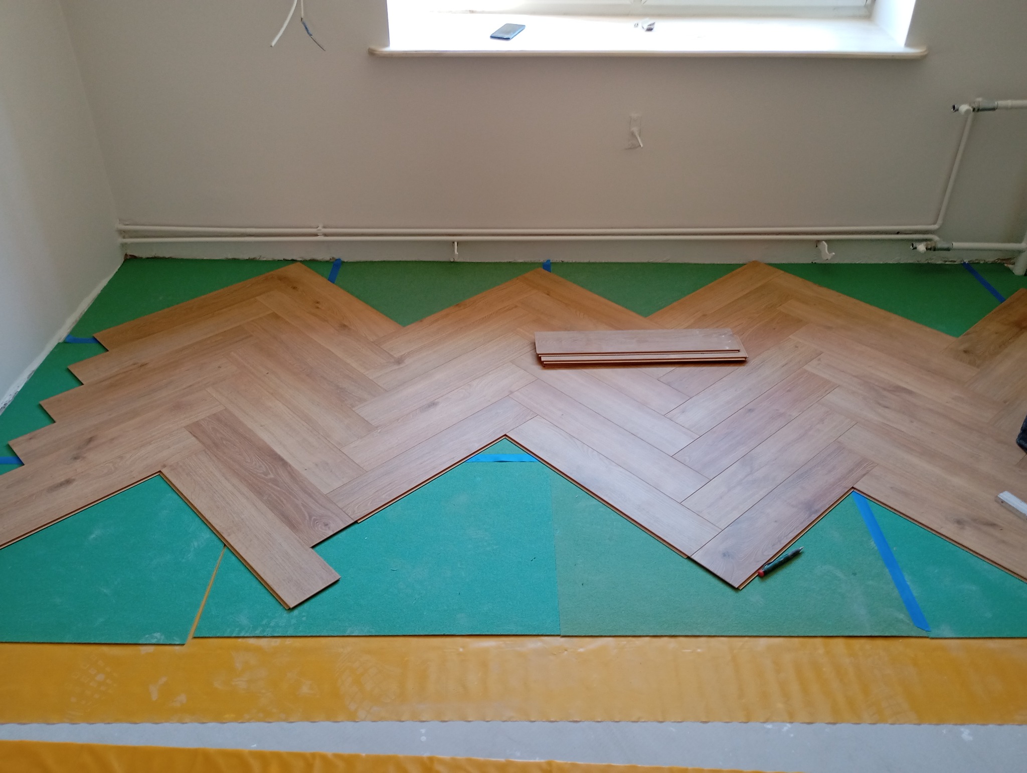 Układanie parkietu w jodełkę na zielonym podkładzie. Widoczne docięte deski, taśma maskująca i narzędzia. Ujęcie z góry, jasne oświetlenie.