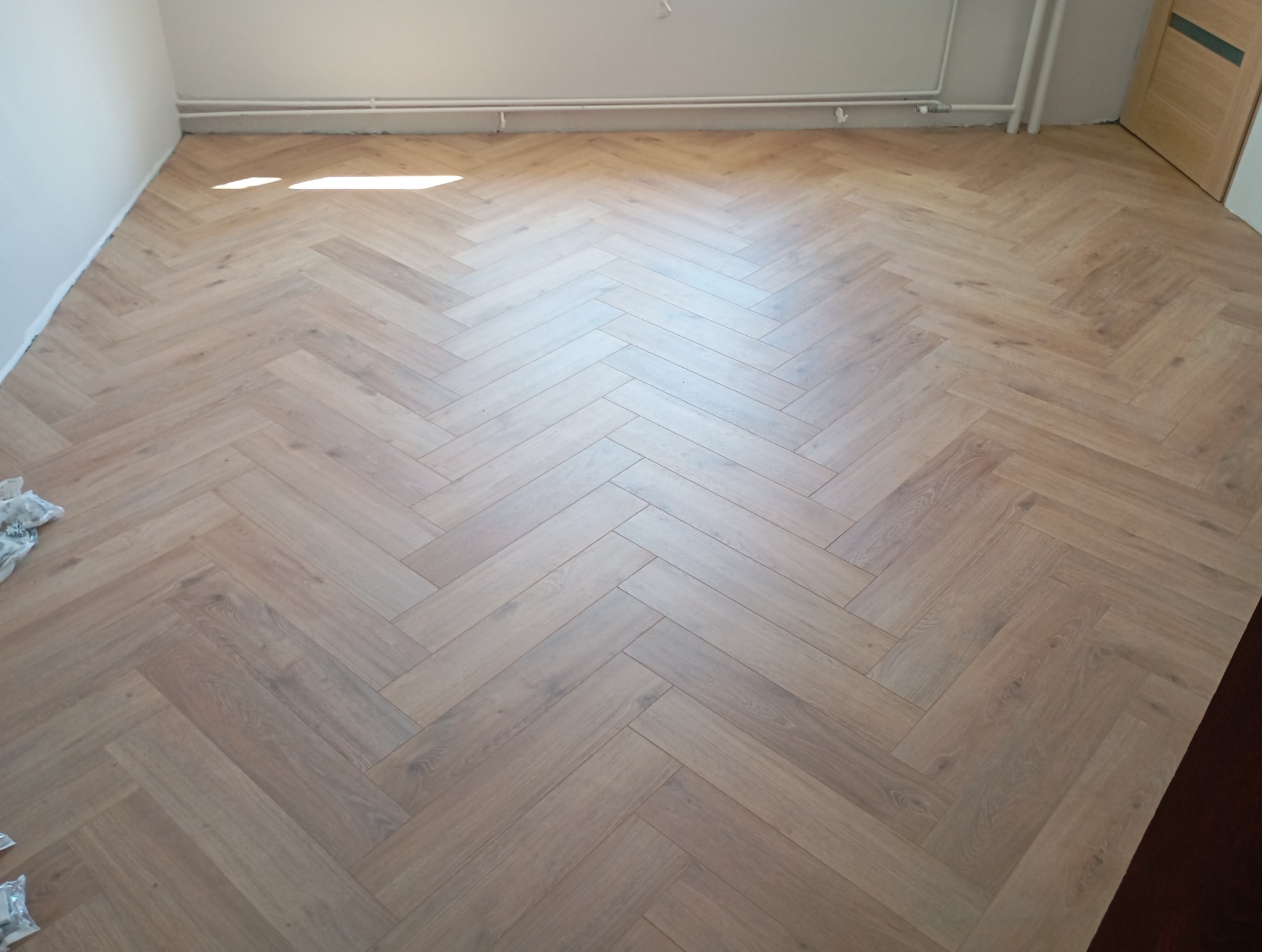 Jasna, drewniana podłoga ułożona w jodełkę francuską, widok z góry. W tle białe ściany i grzejnik. Częściowo widoczna stolarka drzwiowa.