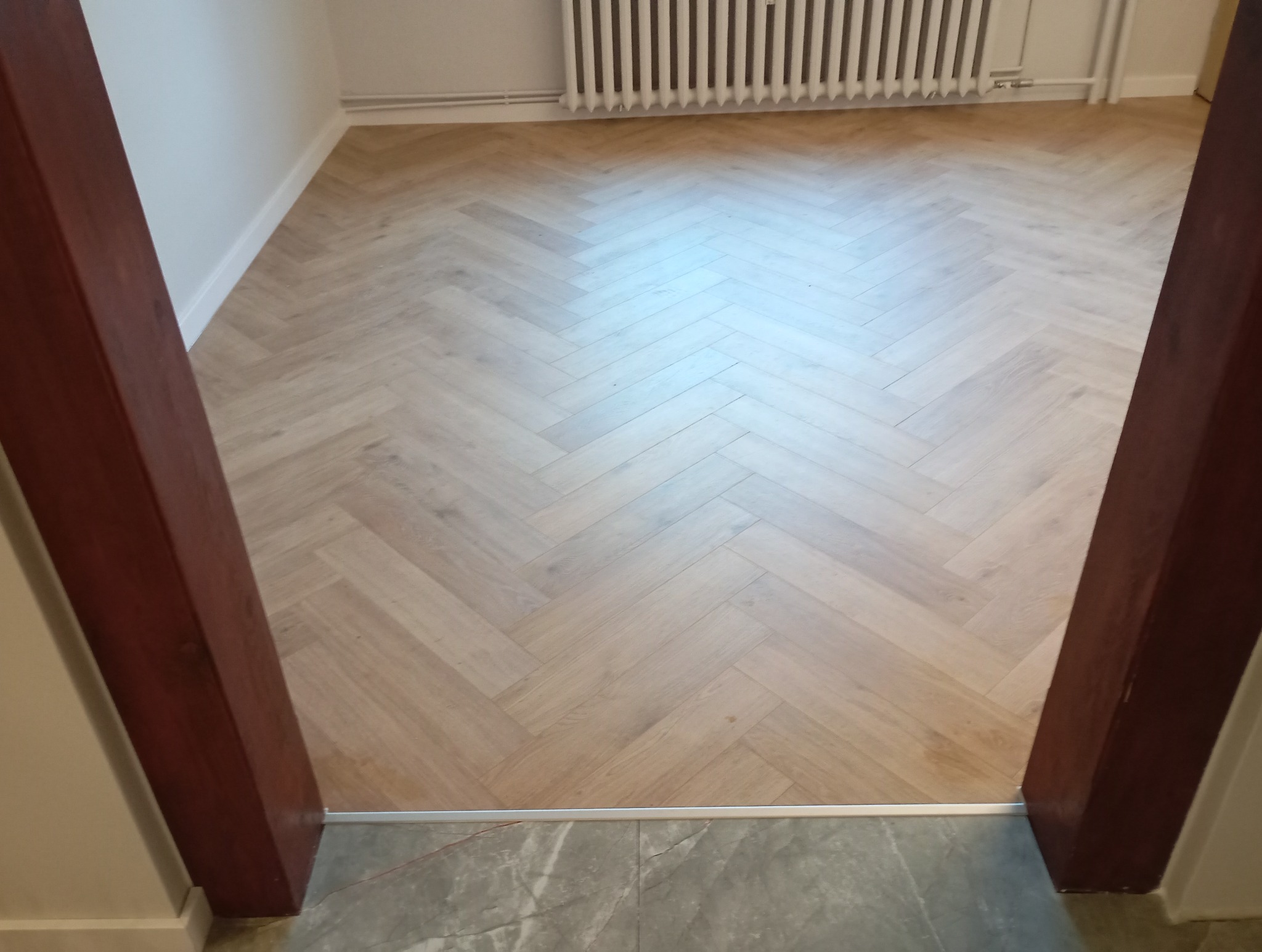 Jasny parkiet ułożony w jodełkę francuską, widoczny z perspektywy progu drzwi. W tle biały grzejnik. Nowoczesne wykończenie podłogi w odcieniach beżu i brązu.