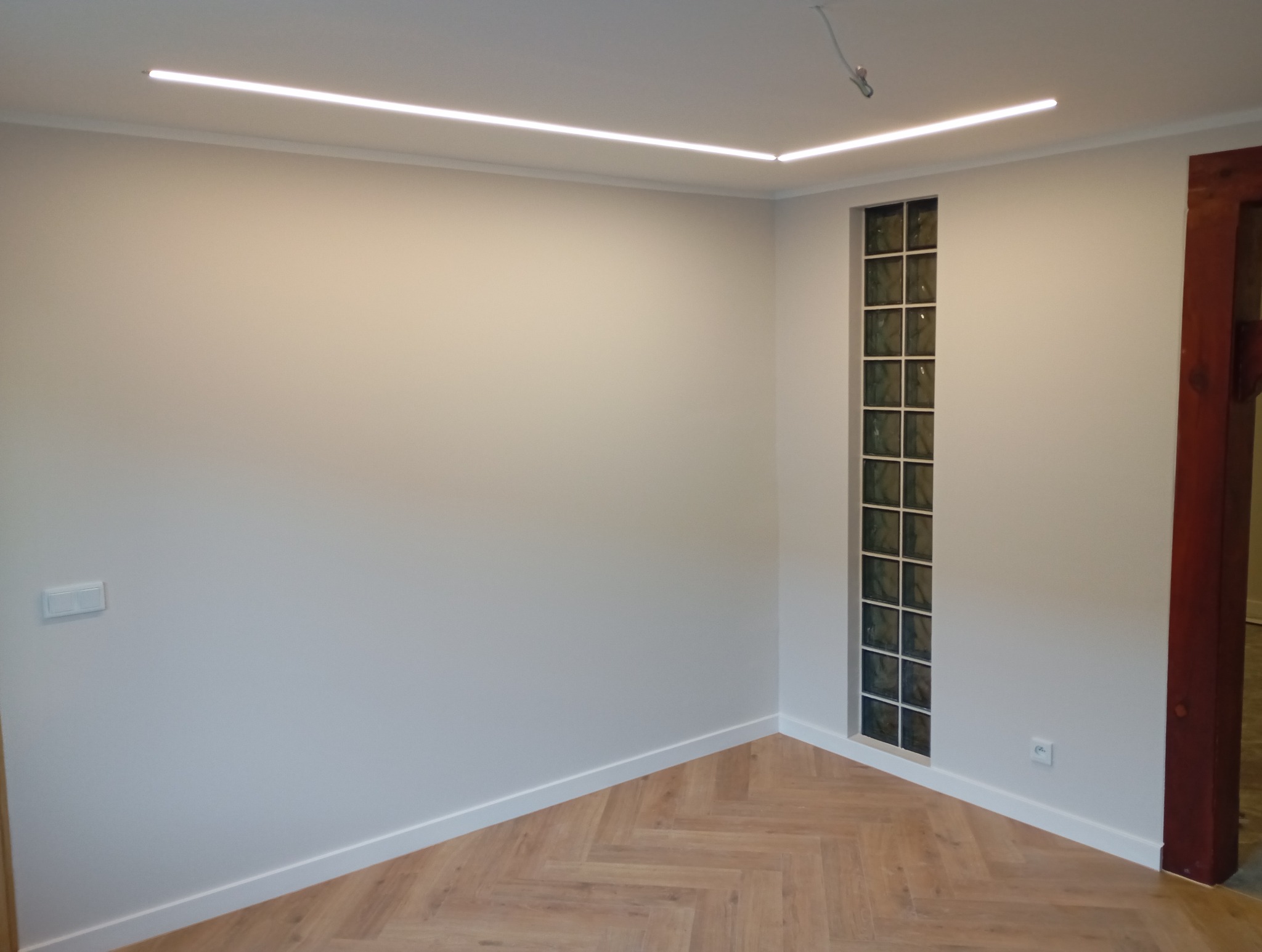 Jasne wnętrze po remoncie: parkiet w jodełkę, szara ściana z oknem z luksferów i listwy przypodłogowe. Oświetlenie LED na suficie.