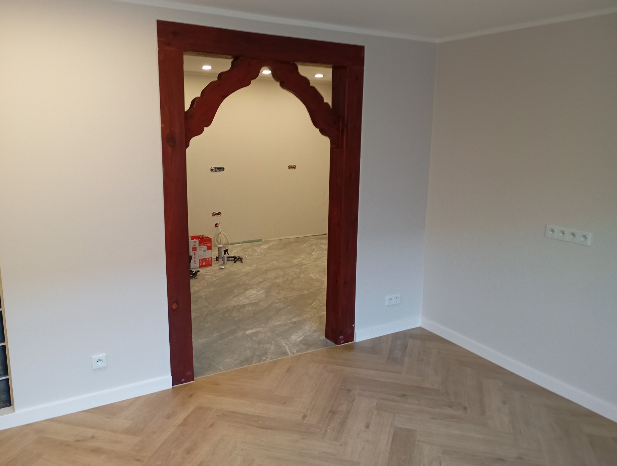 Pokój z drewnianą, zdobioną framugą drzwiową prowadzącą do pomieszczenia z przygotowaną podłogą i instalacją elektryczną. Parkiet ułożony w jodełkę.