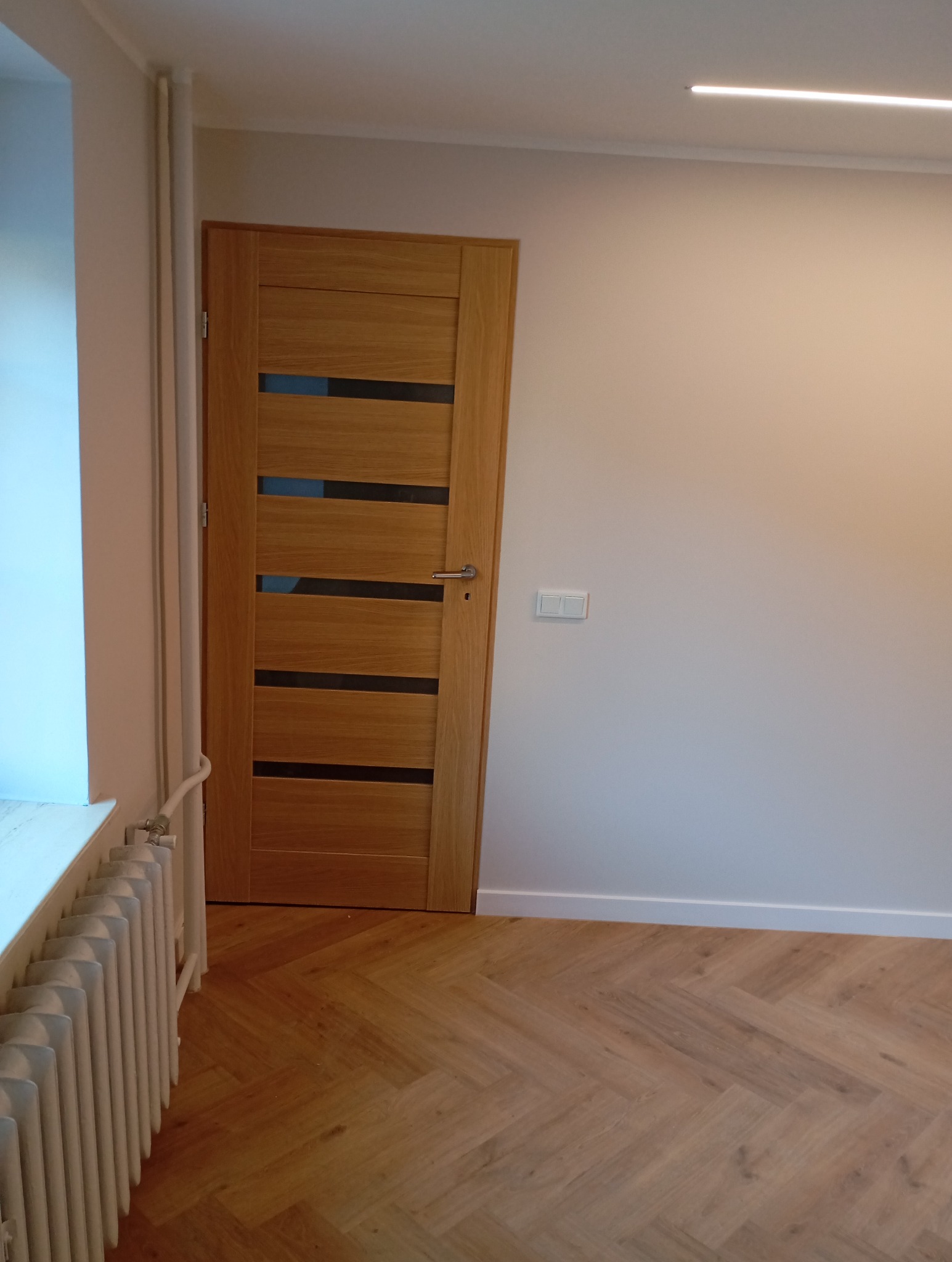 Jasne panele ułożone w jodełkę, drewniane drzwi z przeszkleniami i biała ściana w Urbanowicach. Widoczny grzejnik i listwa przypodłogowa.