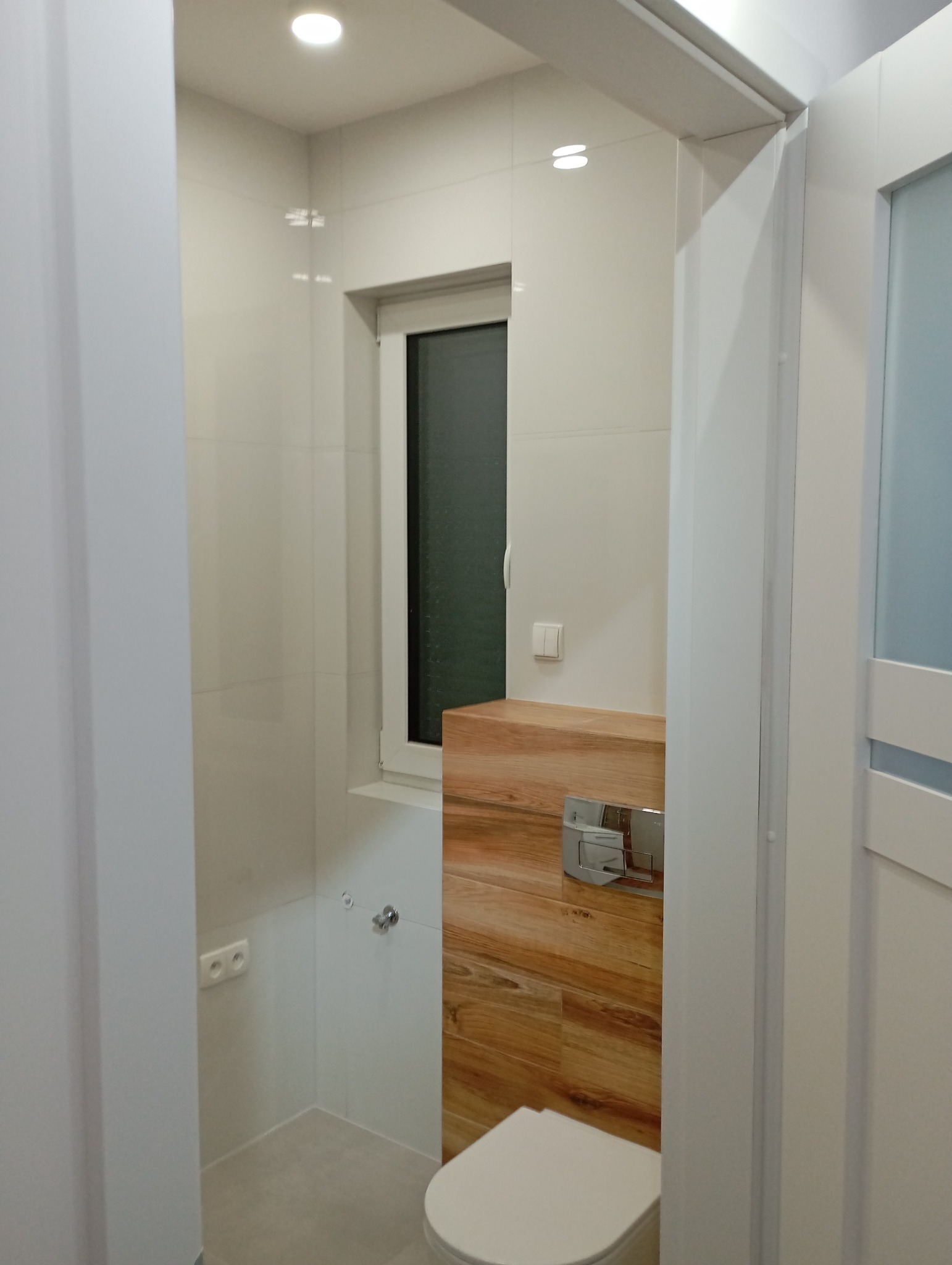 Nowoczesna łazienka z białymi płytkami i akcentem drewnianym. Wisząca toaleta z zabudową. Okno w białej ramie. Widok zza drzwi.