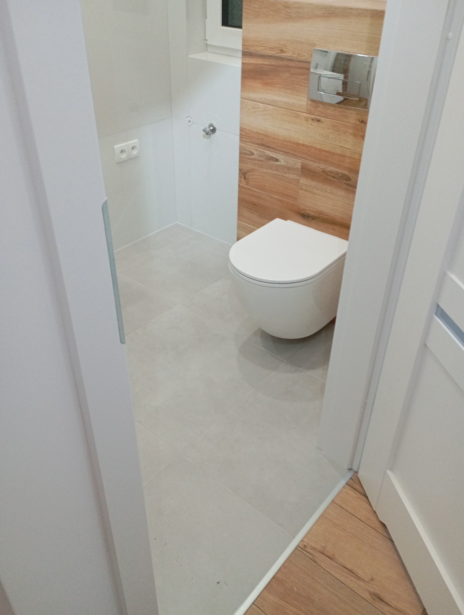 Nowoczesna, wykończona łazienka z podwieszaną toaletą, drewnopodobnymi płytkami na ścianie i jasnymi płytkami podłogowymi. Widok zza uchylonych drzwi.