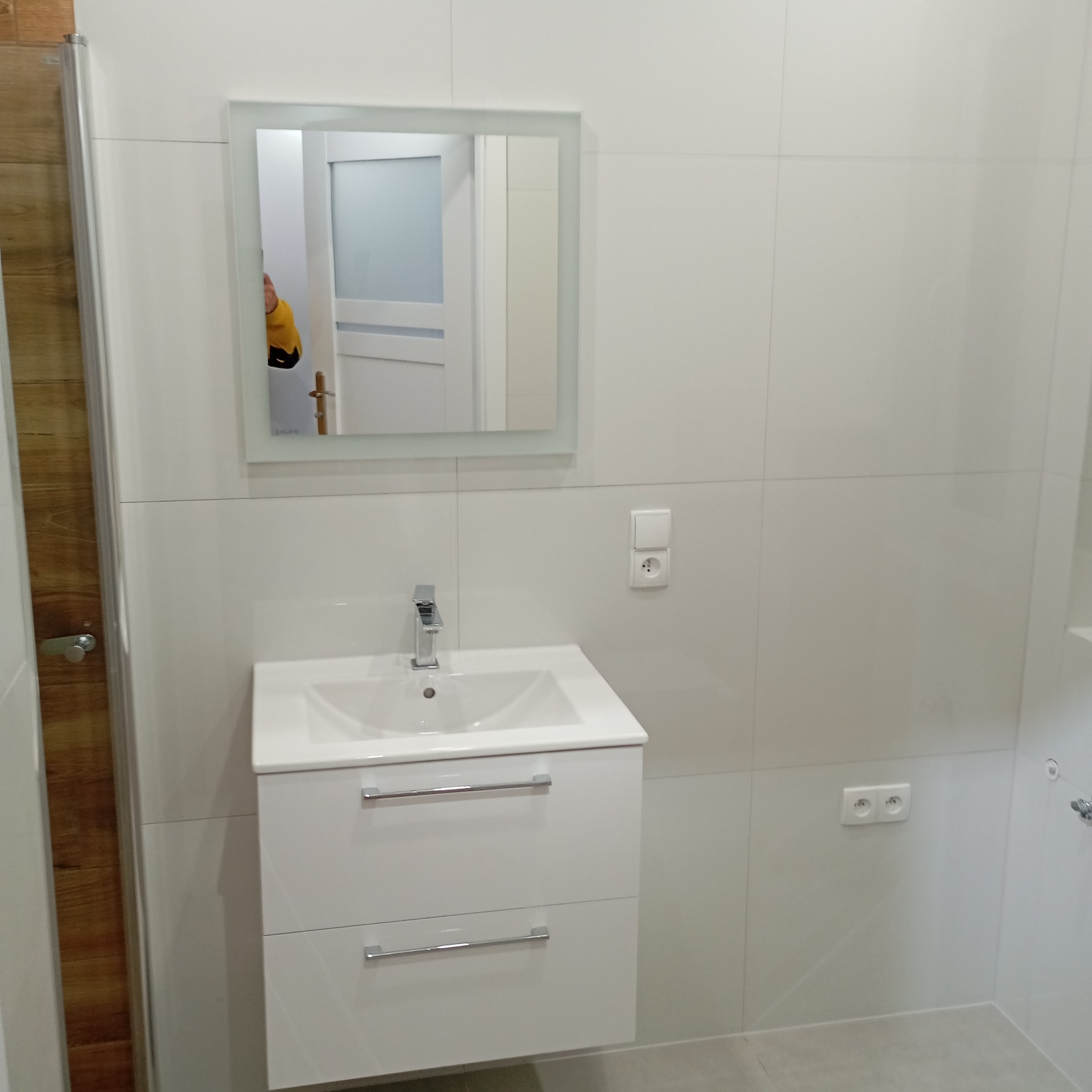 Biała umywalka z szafką i lustrem w nowoczesnej łazience z białymi płytkami. Widoczne gniazdka i fragment kabiny prysznicowej z drewnianym akcentem.