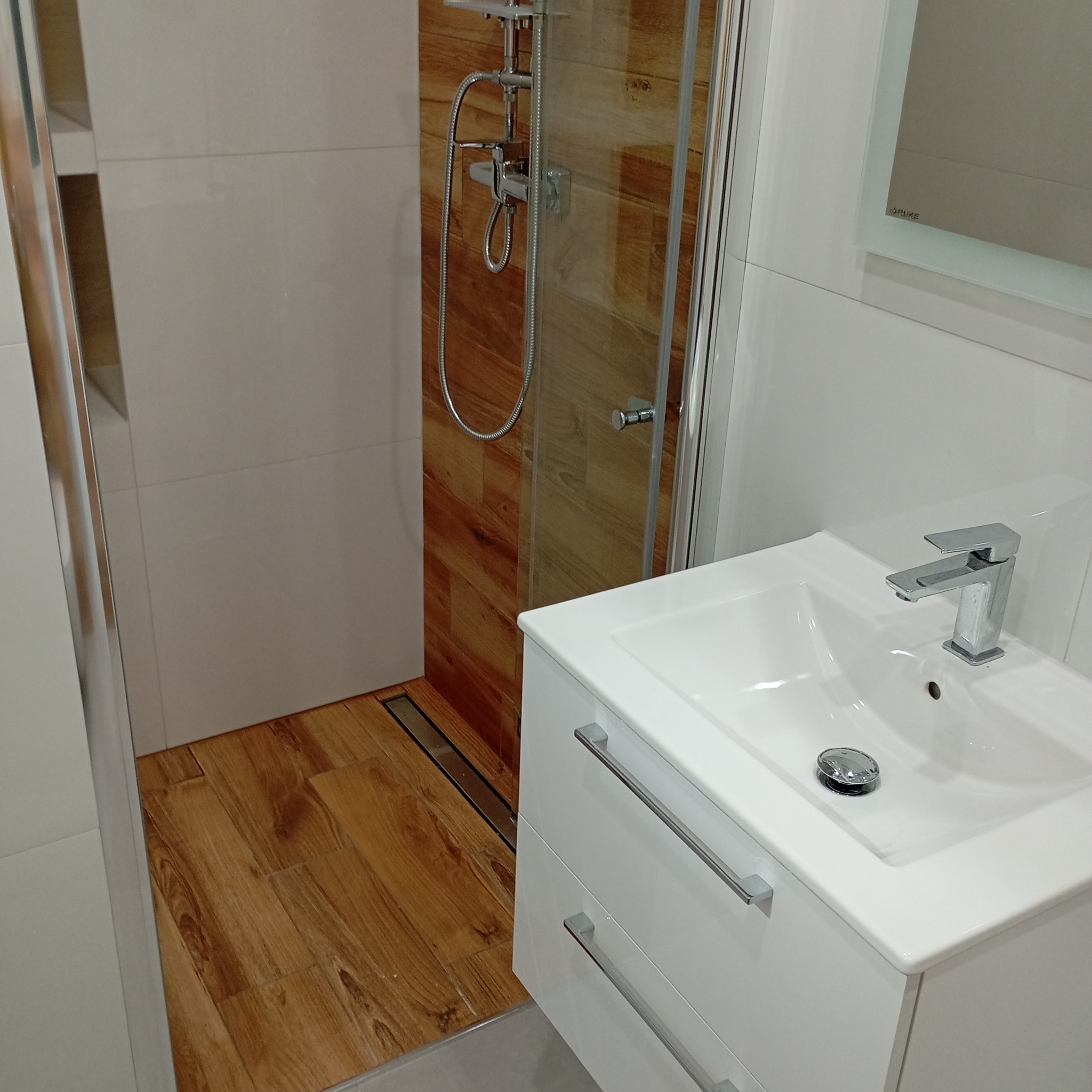 Nowoczesna łazienka z drewnopodobną podłogą i kabiną prysznicową typu walk-in. Biała szafka z umywalką i chromowaną armaturą. Minimalistyczny design, jasne płytki ścienne.