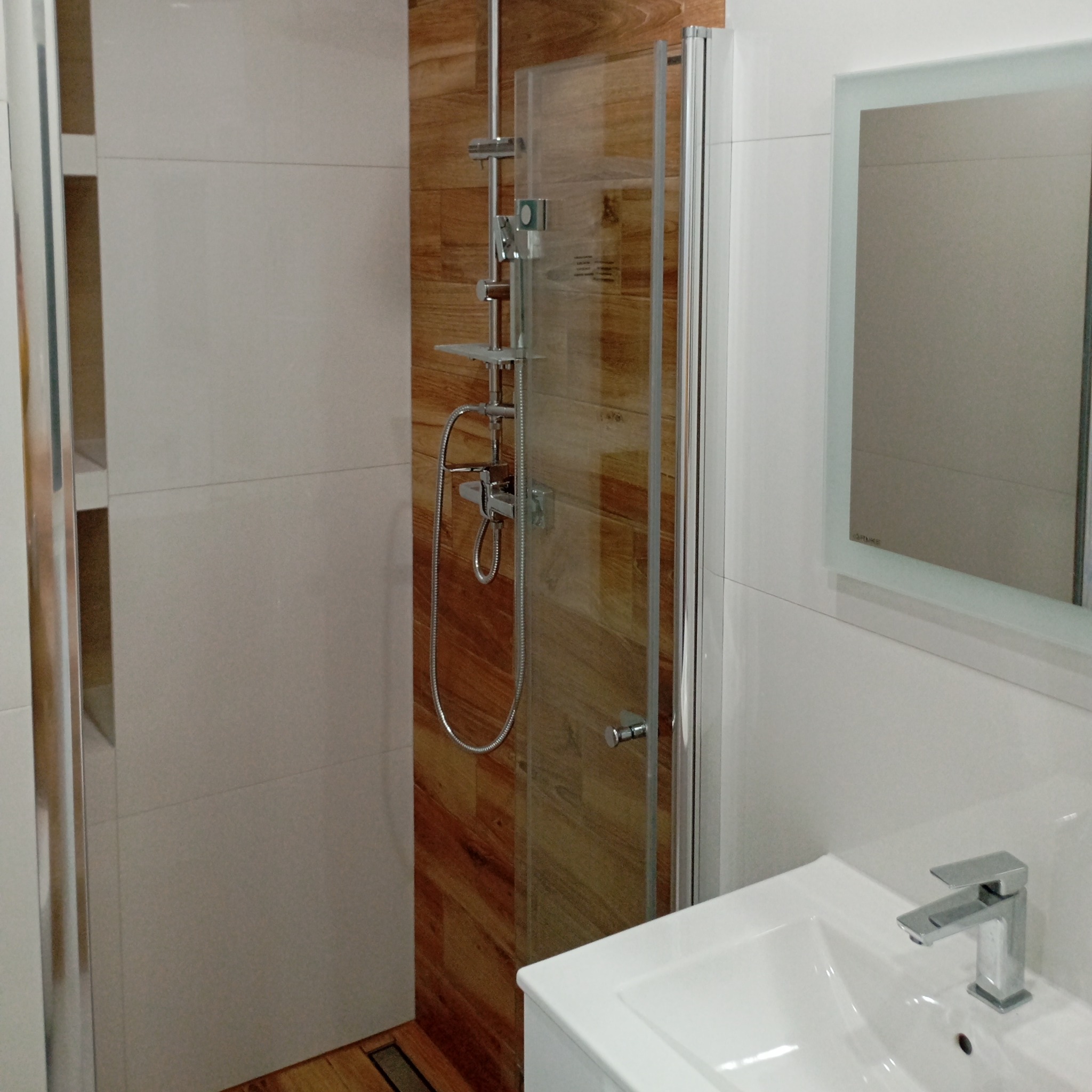 Nowoczesna łazienka z kabiną prysznicową typu walk-in, drewnopodobnymi płytkami i białą umywalką z chromowaną baterią. Lustro z podświetleniem.