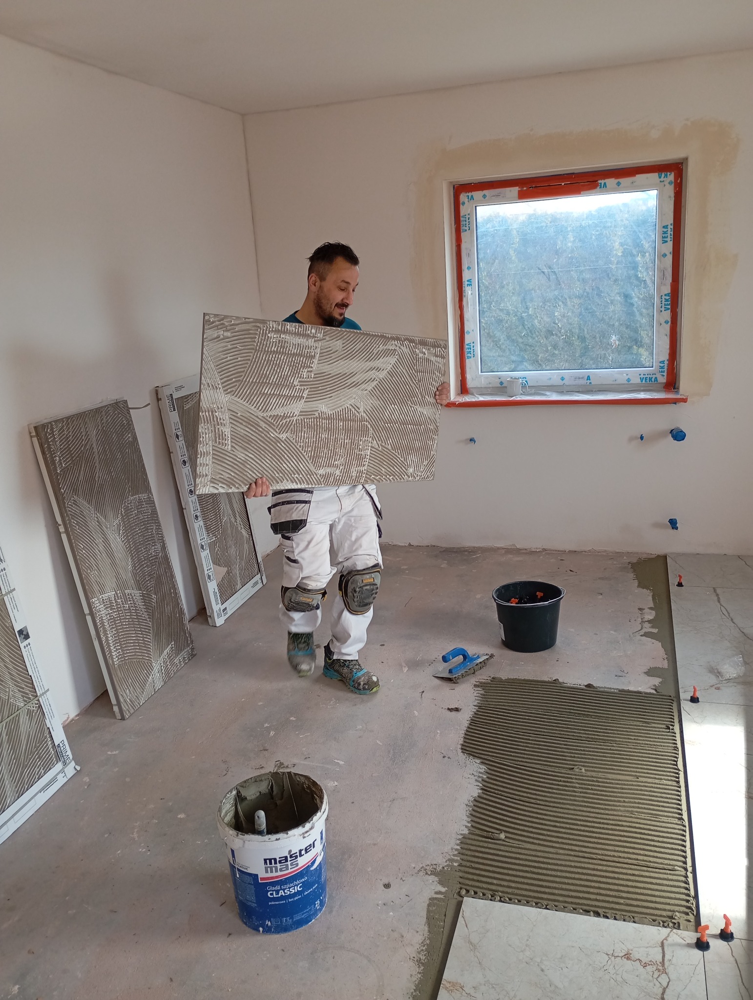 Glazurnik w trakcie pracy: nakłada klej na dużą płytkę, przygotowując się do ułożenia. Widoczne narzędzia, wiadro z klejem i okno z widokiem na zewnątrz.