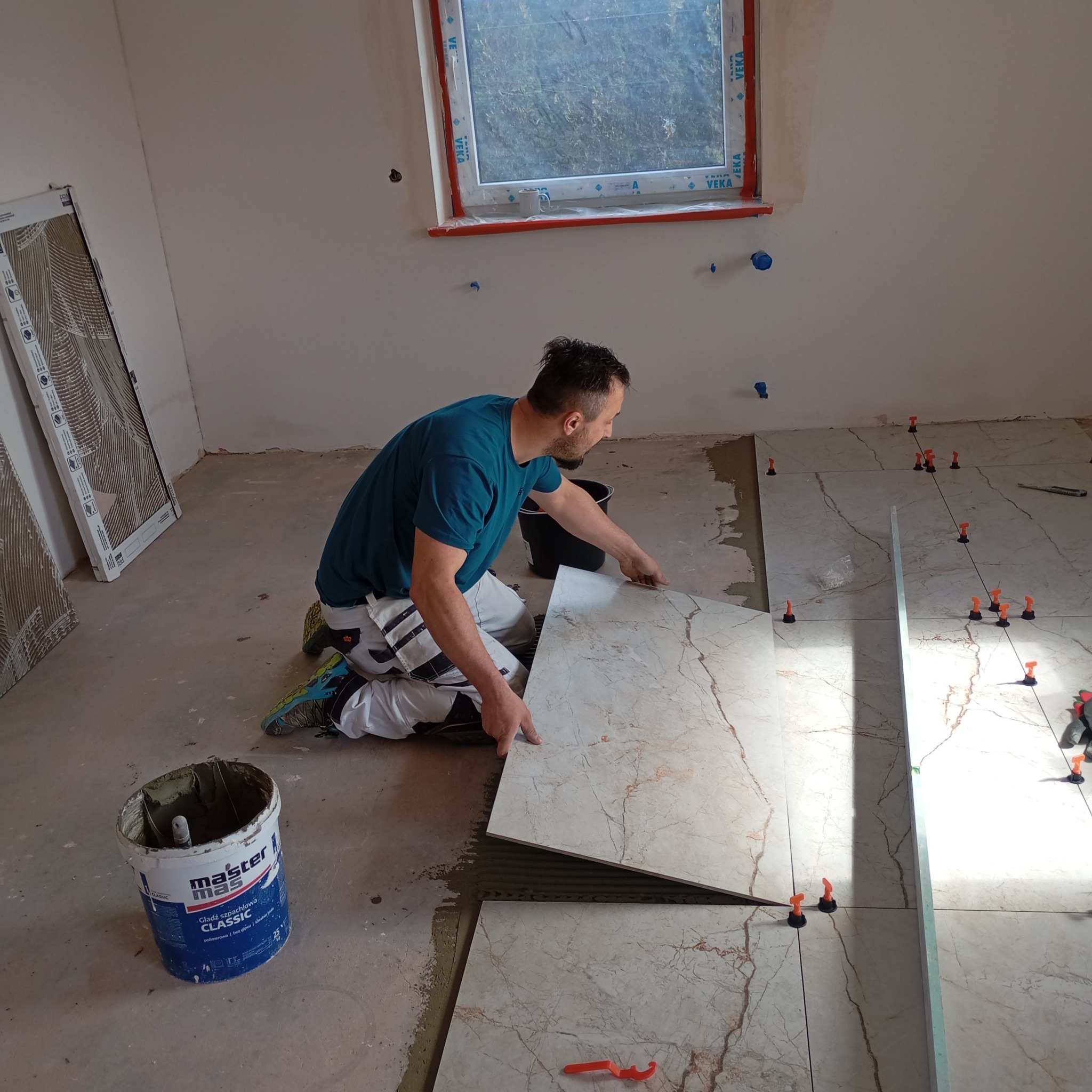 Mężczyzna w trakcie układania płytek podłogowych w jasnym pomieszczeniu. Widoczne narzędzia i poziomica. Płytki imitują marmur. Na ścianie widoczne niebieskie znaczniki.