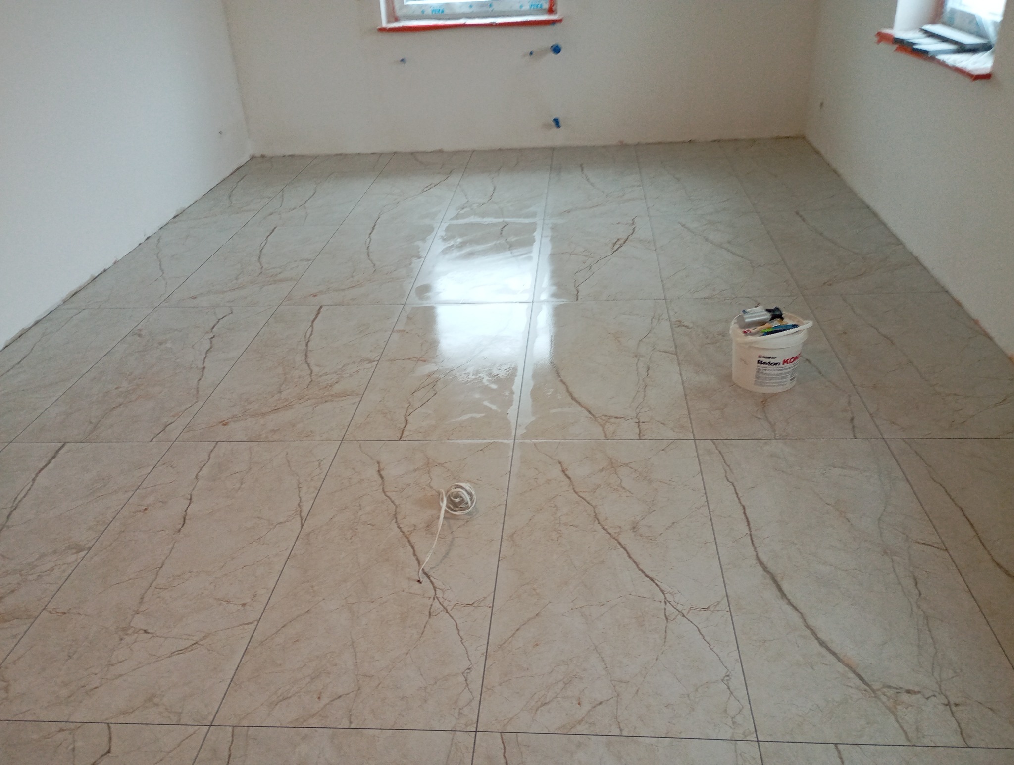 Ułożona podłoga z płytek imitujących marmur w jasnym, pustym pokoju. Wiadro z klejem i zwinięty sznurek na podłodze. Widoczne okno w tle.