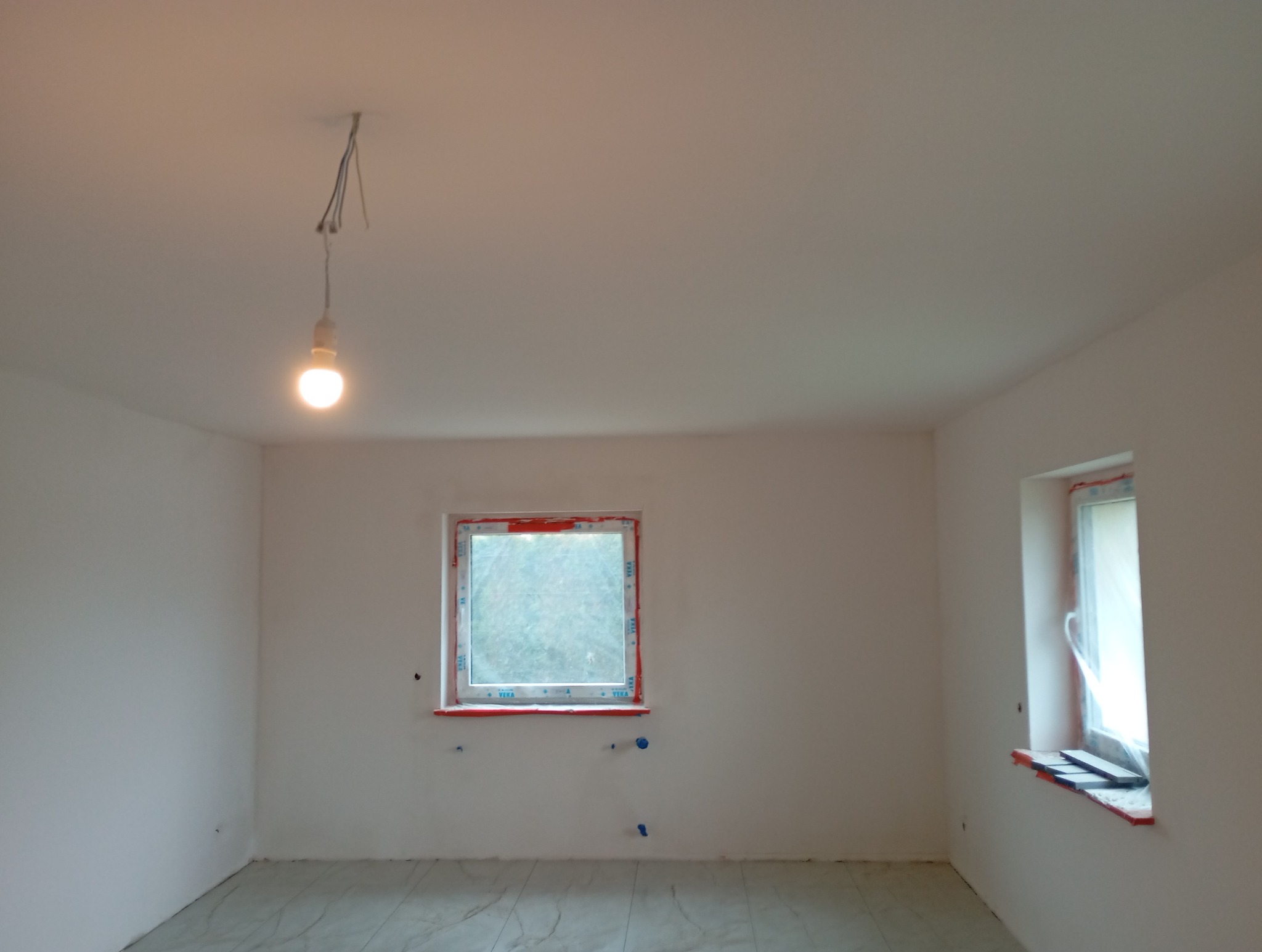 Białe, puste wnętrze po remoncie z dwoma oknami zabezpieczonymi folią i żarówką na kablu. Świeżo położona podłoga z płytek imitujących marmur.