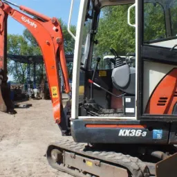Pomarańczowa minikoparka Kubota KX36-3 na gąsienicach, widok z boku na kabinę i ramię z łyżką, w tle inne maszyny budowlane i drzewa.